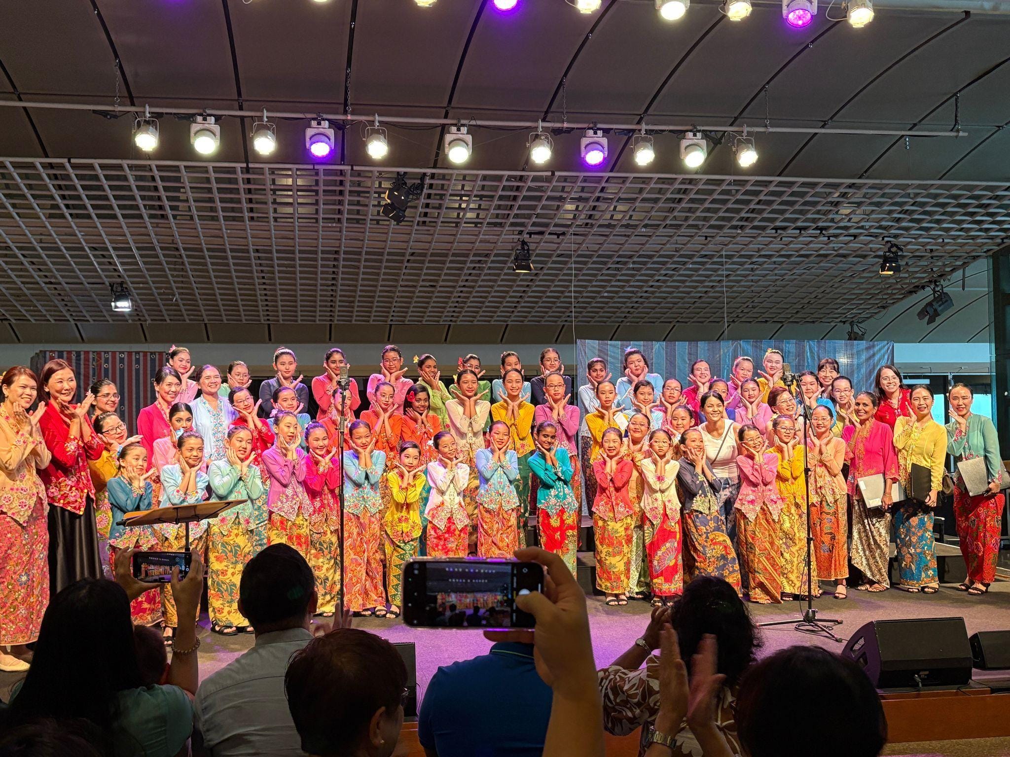 A large group of girls and women in colorful traditional kebaya outfits pose on a lit stage while audience members photograph them.